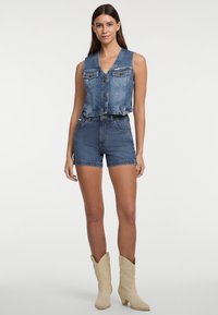 Denim bezrukavni prsluk s gumbima i dva džepa na prsima, uparen s trapericama visokog struka, koje imaju izfrizane rubove, oboje u plavoj boji.