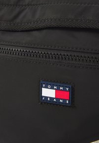 Juodas užtrauktuku uždaromas krepšys su mėlynu, baltu ir raudonu stačiakampiu logotipo pleistru, kuriame užrašyta "TOMMY JEANS" su tekstūruota paviršiumi.