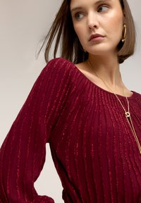 Bordeaux rood geribd shirt met metallic garen accenten, brede halslijn en lange mouwen. De gestructureerde stof zorgt voor een subtiele glans en visuele interesse.