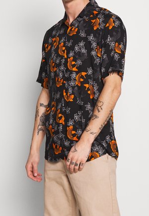 Mann trägt ein kurzärmeliges schwarzes Hemd mit orangenen Koi-Fischen und weißen Blattmustern, beigefarbene Hose und sichtbare Arm-Tattoos.