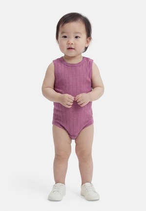 Tout-petit debout portant une combinaison sans manches côtelée violette et des baskets blanches, les mains jointes devant, sur un fond blanc uni.