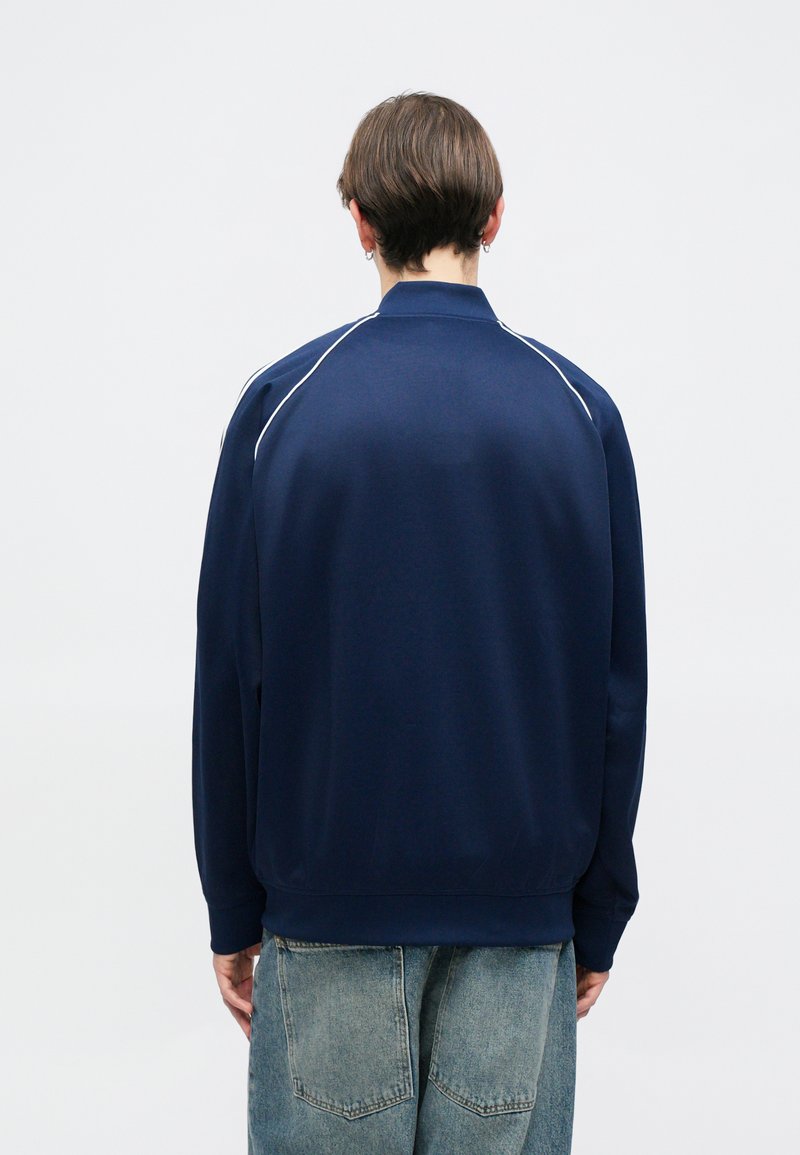 Sweatshirt zippé bleu marine avec des accents blancs en piped, des poignets et un ourlet côtelés. Fabriqué en tissu lisse. Porté avec un jean en denim bleu clair.