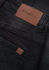 Mustat denim-farkut, joissa on ruskea nahkapatchi, jossa on allekirjoitus. Taskuissa näkyy hienovaraisia tekstuuri- ja ompeluyksityiskohtia.