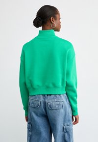 Sweatshirt verde cropped com gola alta, apresentando uma textura suave e punhos canelados, combinado com jeans cargo azul claro.