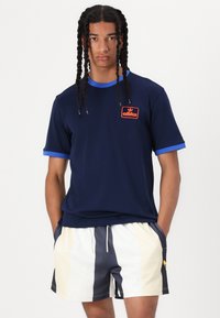 Mörkblå t-shirt med en blå krage och röd broderad logotyp, tillsammans med vita och mörkblå randiga meshshorts med justerbar midja.