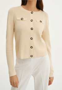 Beżowy kardigan w prążki z czarnymi i złotymi guzikami, dwiema klapkami kieszeni oraz długimi rękawami, uzupełniony przez krótki fason.