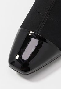 Chaussure en tissu noir avec un embout en cuir verni noir brillant, présentant des détails de couture subtils sur une surface supérieure lisse.
