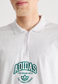 Grå polo skjorta med dragkedja vid kragen, med grön broderad text och logotyp. Mjuk tygkvalitet med en avslappnad passform och subtil struktur.