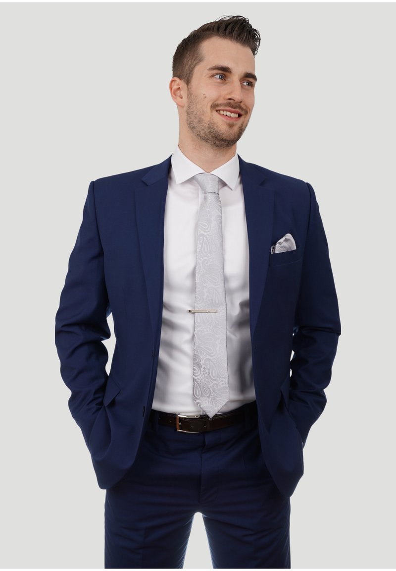 Traje azul marino con una camisa blanca, corbata plateada con patrón y un pañuelo a juego. Presenta un corte ajustado y un cinturón con hebilla plateada.