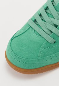 Gros plan sur l'avant d'une basket en daim bleu sarcelle avec des lacets assortis et une semelle en caoutchouc marron sur fond blanc.