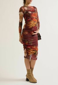 Een zwangere vrouw poseert in een bloemenprint, strak aansluitende jurk, terwijl ze haar buik vasthoudt, gecombineerd met tan enkellaarzen en een kleine handtas.