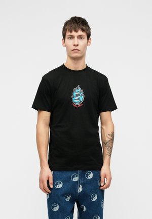 Fiatal férfi, rövid barna hajjal, fekete pólót visel, amelyen kék lángoló szájlogó található, és kék farmernadrágot, amelyen fehér yin-yang szimbólumok vannak nyomtatva.