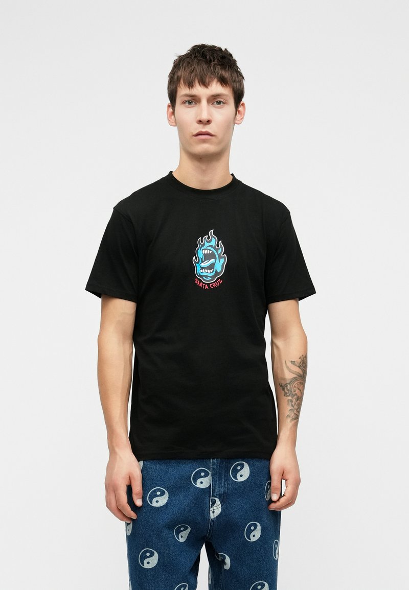 Νεαρός άνδρας με κοντά καστανά μαλλιά φοράει μαύρο Τ-shirt με μπλε λογότυπο φλογισμένου στόματος και μπλε τζιν με λευκά σύμβολα γιν-γιανγκ.