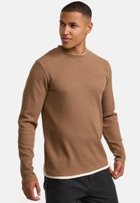 Braunes, geripptes Langarmshirt mit rundem Halsausschnitt. Weiche Textur, taillierte Passform und dezentes Logodetail auf der Brust. Kombiniert mit dunklen Jeans.