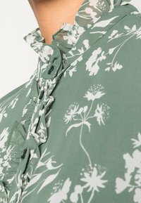 Blusa de estampado floral verde con cuello rizado y cierre de botones, confeccionada en una tela ligera con textura sutil.
