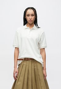 Vit pikétröja med korta ärmar och en krage med tre knappar, kombinerad med en beige plisserad kjol och ett brunt bälte.