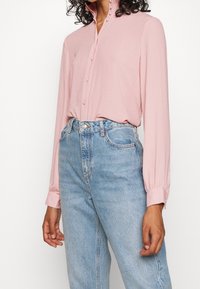 Blusa rosa chiaro a maniche lunghe con colletto alto e chiusura a bottoni, abbinata a jeans in denim blu a vita alta. Tessuto morbido.