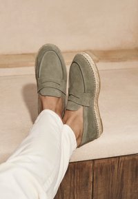 Suede loafers in muted green with stitched accents and a textured espadrille-style sole. Paired with white pants for contrast.