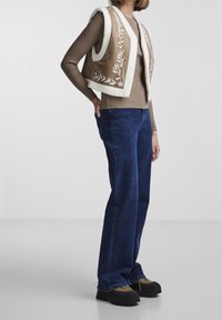 Person i en brun ribbad tröja, suèdeväst med vit broderi och fårskinnsdetaljer, mörkblå jeans och bruna platåskor.