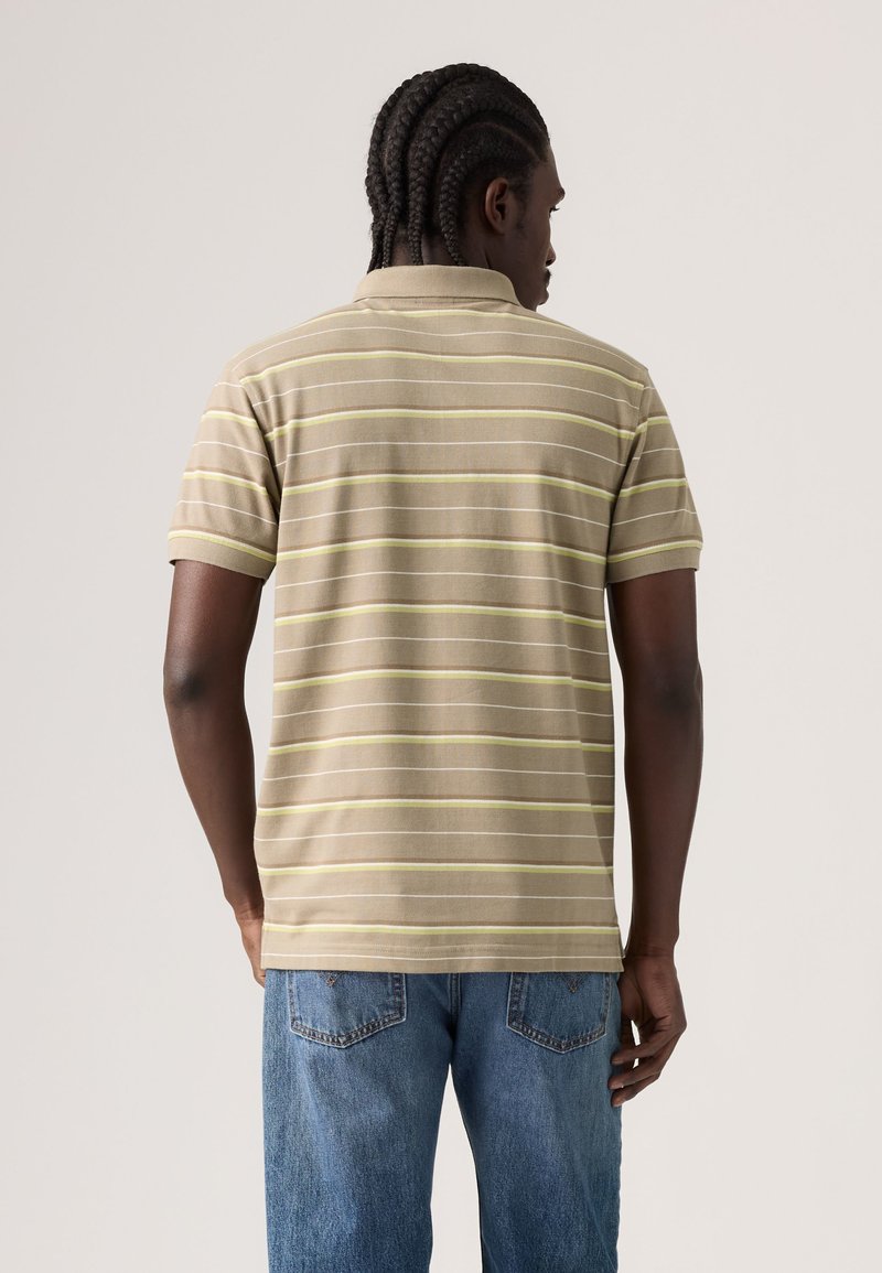 Man with braided hair wearing a beige and light yellow striped polo shirt and blue jeans, facing away against a plain background.