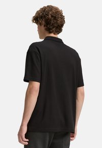 Fekete pólóing textúrált szövettel, rövid ujjakkal és klasszikus gallérral. Lazább fazonú, és minimális logó található az oldalon.
