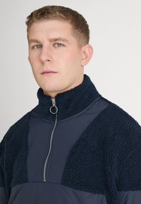 Marineblauw fleece pullover met half-rits, textuur op de bovenste panelen, gladde onderste stof en een hoge kraag. Zilveren ritssluitingen en ringtrek detail.