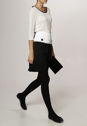 Haut blanc à manches trois-quarts avec des accents noirs, associé à une jupe noire et des collants noirs. Tient une pochette noire, avec un simple détail de bouton sur la jupe.