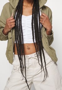 Olive green zip-up jacket with a hood, white ruched crop top, and light beige cargo pants. Long braided hair and a navel piercing are visible.