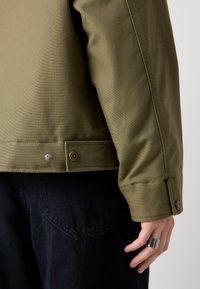 Veste en vert olive, texturée avec une coupe droite. Présente des boutons pressions et des détails aux poignets, associée à un pantalon en denim foncé. Main visible avec une bague.