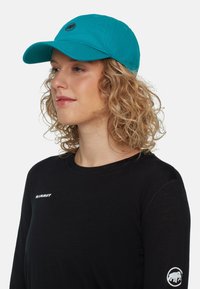 Een turquoise baseballpet van katoen, met een rond logo aan de voorkant, gecombineerd met een zwarte longsleeve shirt met witte branding aan de zijkant.
