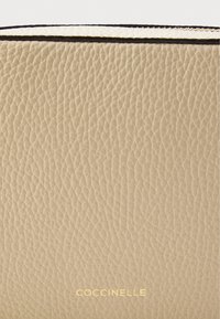 Light beige leather surface with a textured finish and embossed brand logo in gold at the bottom. Rounded edges and black seam detail.
