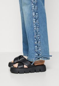 Sandalias de plataforma negras con un diseño de tiras cruzadas, combinadas con vaqueros azul claro adornados con un detalle decorativo de cordones a lo largo del costado.