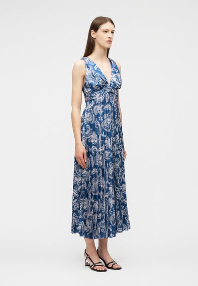 Femme debout portant une robe maxi sans manches bleue et blanche à motifs floraux avec un décolleté en V et des sandales à talons noires à brides.