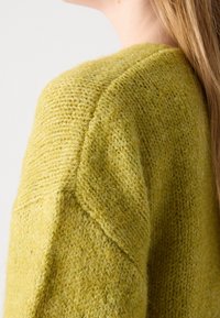 Gros plan sur une personne portant un pull en maille vert olive texturé et duveteux, montrant l'épaule et la partie supérieure de la manche.