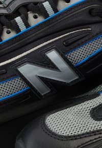 Sorte sportsko med grå mesh og blå accenter. Har et fremtrædende sølv New Balance-logo, teksturerede materialer og snøresystem.