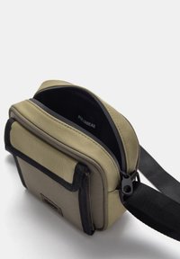 Rechteckige Schultertasche aus olivgrünem Stoff mit schwarzen Akzenten, ausgestattet mit einem Reißverschlussöffnung, einem verstellbaren Tragegurt und einem markierten Innenetikett.