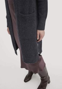 Dunkelgrauer gestrickter Cardigan mit zwei vorderen Taschen, getragen über einem lila Satin-Kleid mit gewelltem Saum, kombiniert mit braunen kniehohen Stiefeln.