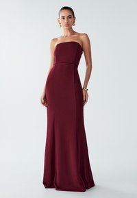 Žena v dlouhé bordó gown bez ramínek s upnutým korzetem, doplněná zlatými kruhovými náušnicemi a náramkem na jednobarevném pozadí.