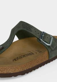 Κοντινή λήψη πράσινου σουέντ σανδαλιού Birkenstock με ρυθμιζόμενη μεταλλική πόρπη και ελαφρώς διαμορφωμένη φαρδιά φτέρνα από φελλό, μέγεθος 42, σε λευκό φόντο.