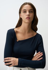 Maglietta a maniche lunghe blu navy a coste con scollatura asimmetrica e dettagli argentati. Vestibilità aderente, tessuto morbido, design minimalista.