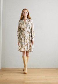 Rochie cu mânecă lungă din material ușor, cu un model de frunze, având o talie accentuată cu curea. Asortată cu cizme bej cu genunchi pe un pardoseală din lemn.