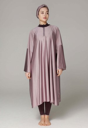 Femme portant une robe mauve ample à manches longues avec un foulard assorti et des leggings foncés, debout pieds nus sur un fond uni.