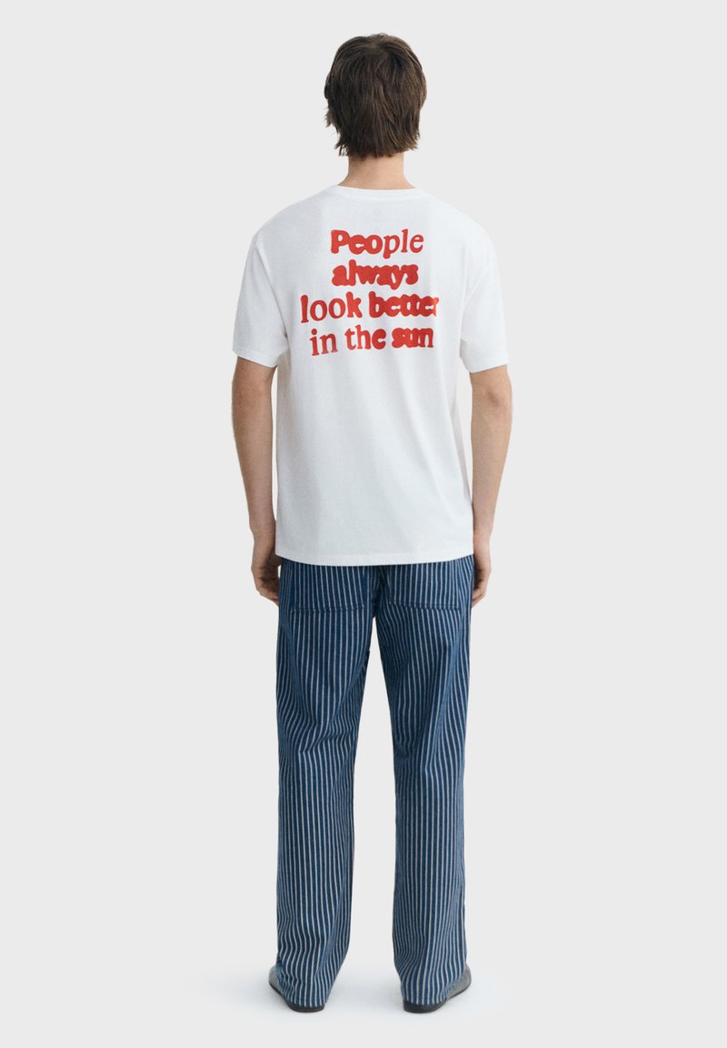 Mann trägt weißes T-Shirt mit rotem Text "People always look better in the sun" und blau gestreifte Hose, von hinten auf einfarbigem Hintergrund.