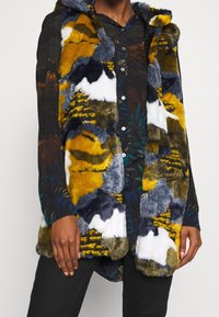Chaleco de piel sintética con una mezcla de parches amarillos, grises y blancos, con capucha y diseño de frente abierto. Lleva encima una camisa estampada.