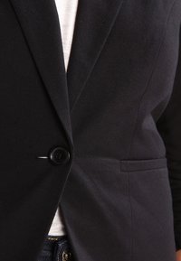 Czarna marynarka z pojedynczym zapięciem na guzik, wąskimi klapami i bocznymi kieszeniami. Wykonana z miękkiego materiału o gładkiej fakturze.