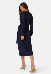 Granatowa sukienka plisowana z długimi rękawami i paskiem w talii, o długości do połowy łydki i gładkiej teksturze. Noszona z beżowymi szpilkami.