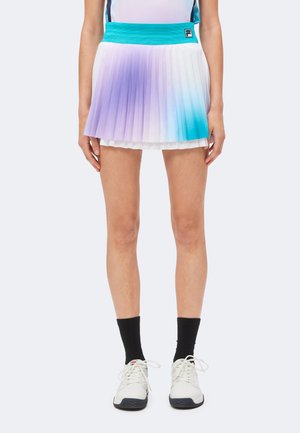 Vrouw die een geplooide tennisrok draagt met paarse en blauwe verloopkleuren, witte sneakers en zwarte sokken, staand tegen een effen achtergrond.