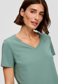 Koszulka z krótkim rękawem i dekoltem w serek w miękkiej, miętowej tkaninie o gładkiej fakturze i luźnym kroju, z subtelnymi akcentami na ramionach.