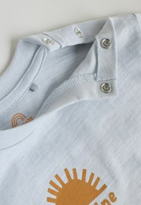 Body bébé bleu clair avec boutons-pression argentés le long de la couture de l'épaule et un motif soleil orange partiellement visible.