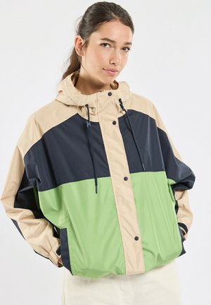 Jeune femme portant une veste coupe-vent à capuche beige, noire et verte avec les mains dans les poches.